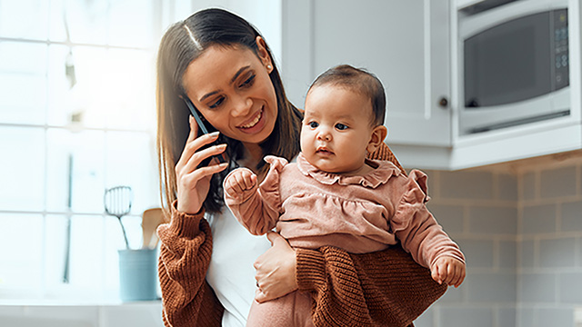 A woman holding a baby is talking on a phone in a home kitchen.