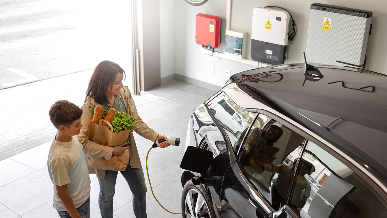 Happy son and mother plug EV charger from home charging station to electric vehicle. Future eco-friendly car powered by renewable source of clean energy on daytime. Horizontal, high angle.