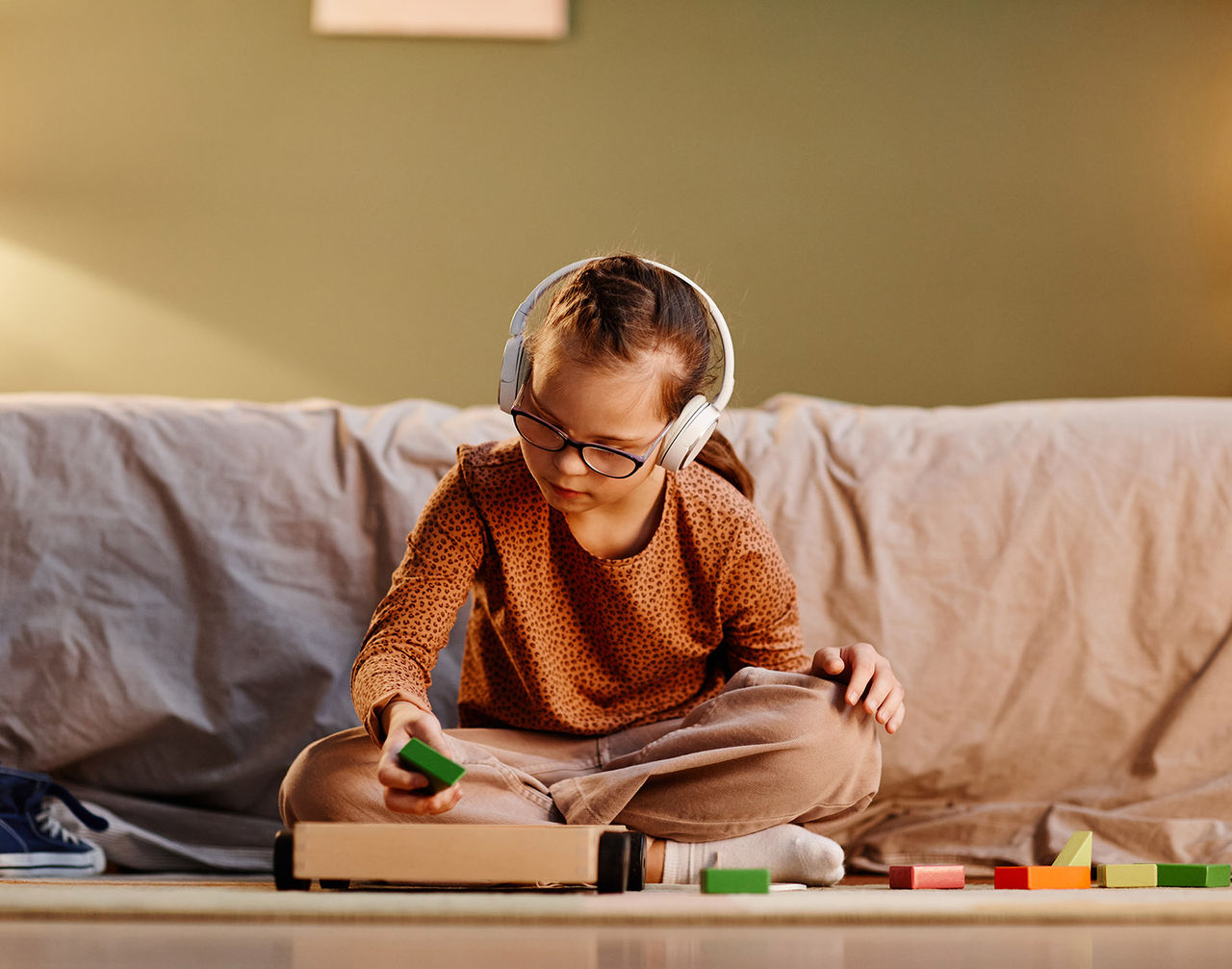 A young child wearing large white headphones sits cross-legged on a couch. The child is playing with colorful wooden blocks, holding a green block in one hand.
