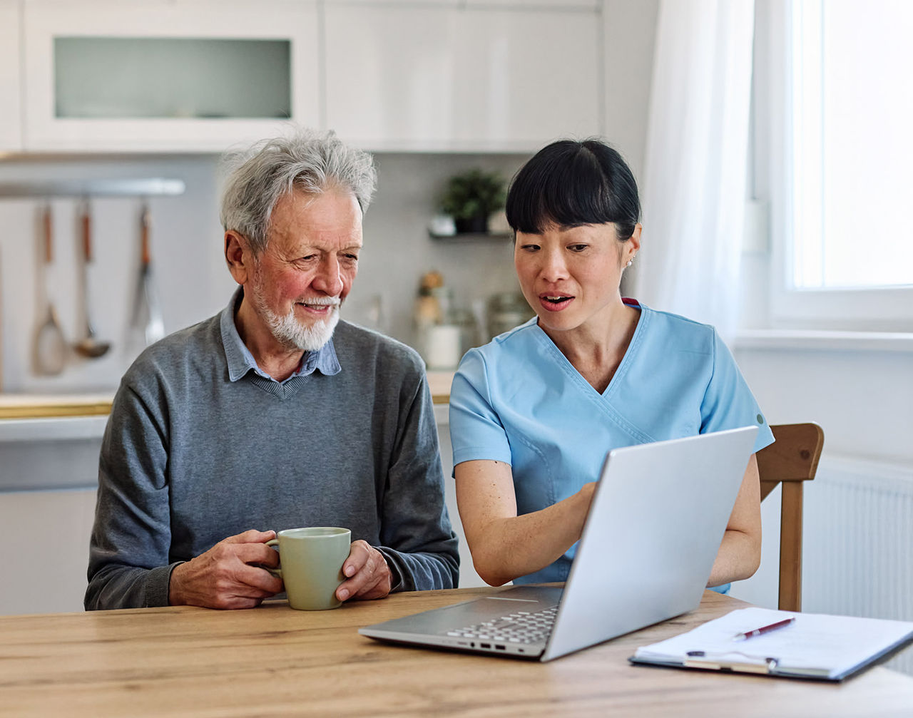 A senior man sits at a wooden table holding a mug while looking at a laptop screen. Next to him, a healthcare worker in light blue scrubs gestures toward the computer, explaining something.