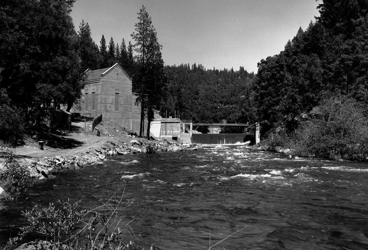 A black and white photograph of a fast-flowing river running through a forested valley, passing through West Point Powerhouse on the rocky bank. Tall trees surround.