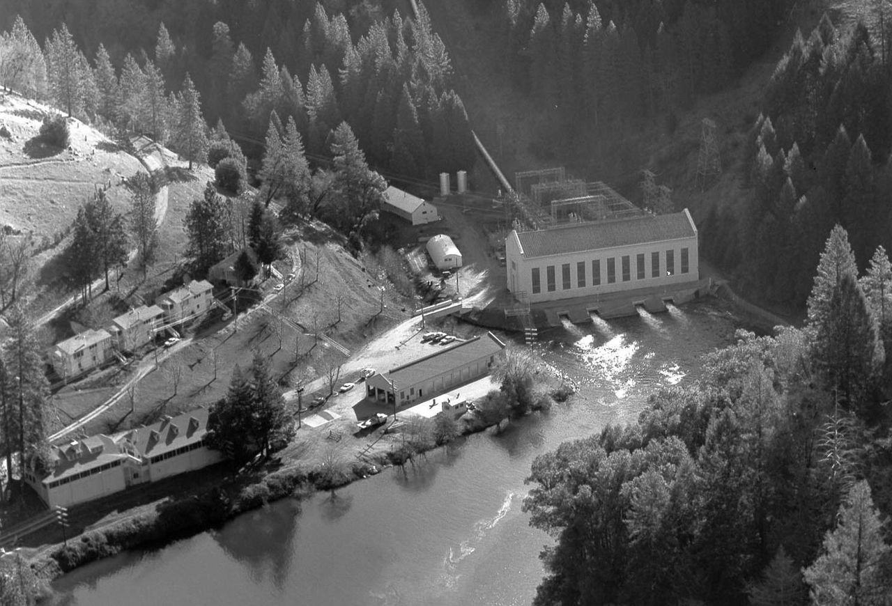 An aerial black and white photograph shows the Tiger Creek Powerhouse situated along a river in a forested, mountainous area. The main generating station stands beside the water with several outflow channels leading into the river. Nearby, smaller industrial buildings and residential houses are clustered along the hillside. Dense trees surround the facility.
