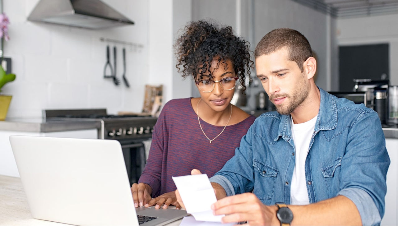 ਇੱਕ ਰਸੋਈ ਵਿੱਚ ਦੋ ਲੋਕ ਕਾਗਜ਼ ਦੇ ਇੱਕ ਟੁਕੜੇ ਅਤੇ ਇੱਕ ਲੈਪਟਾਪ ਵੱਲ ਦੇਖ ਰਹੇ ਹਨ