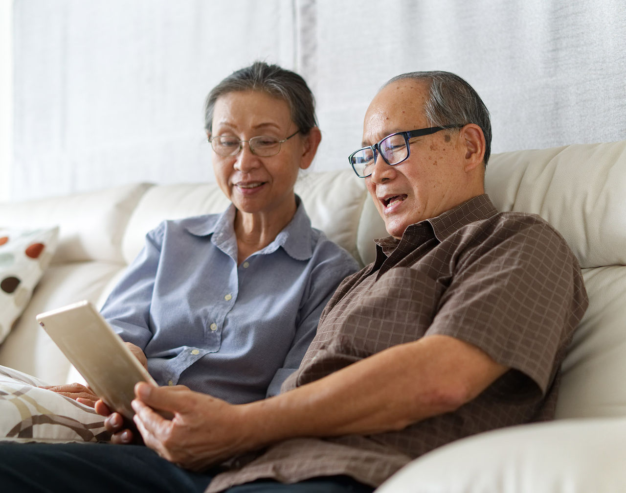 An older couple sits together on a light-colored sofa, looking at a digital tablet in the man's hands. They appear relaxed and engaged, sharing the screen as they browse or read.