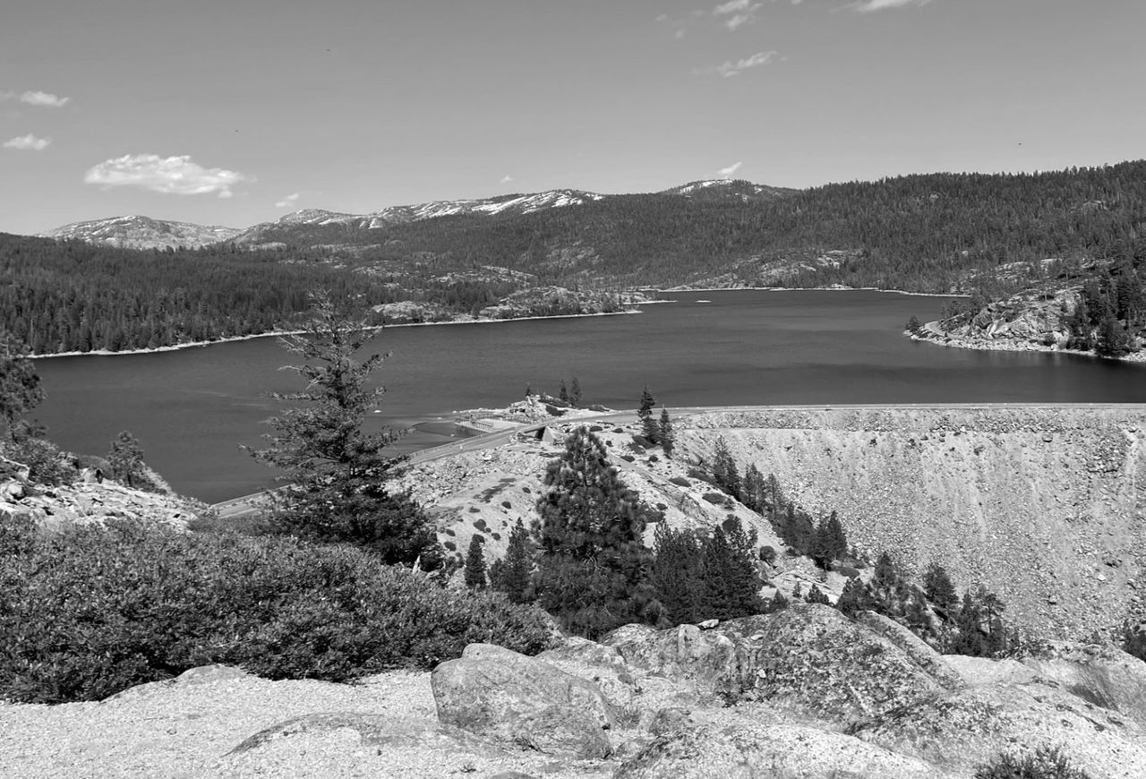 A black and white photograph of Lower Bear Reservoir Dam. The image is captured from an elevated viewpoint, looking down over shrubs and boulders toward the water. Snow-capped peaks rise in the distant background under a clear sky.