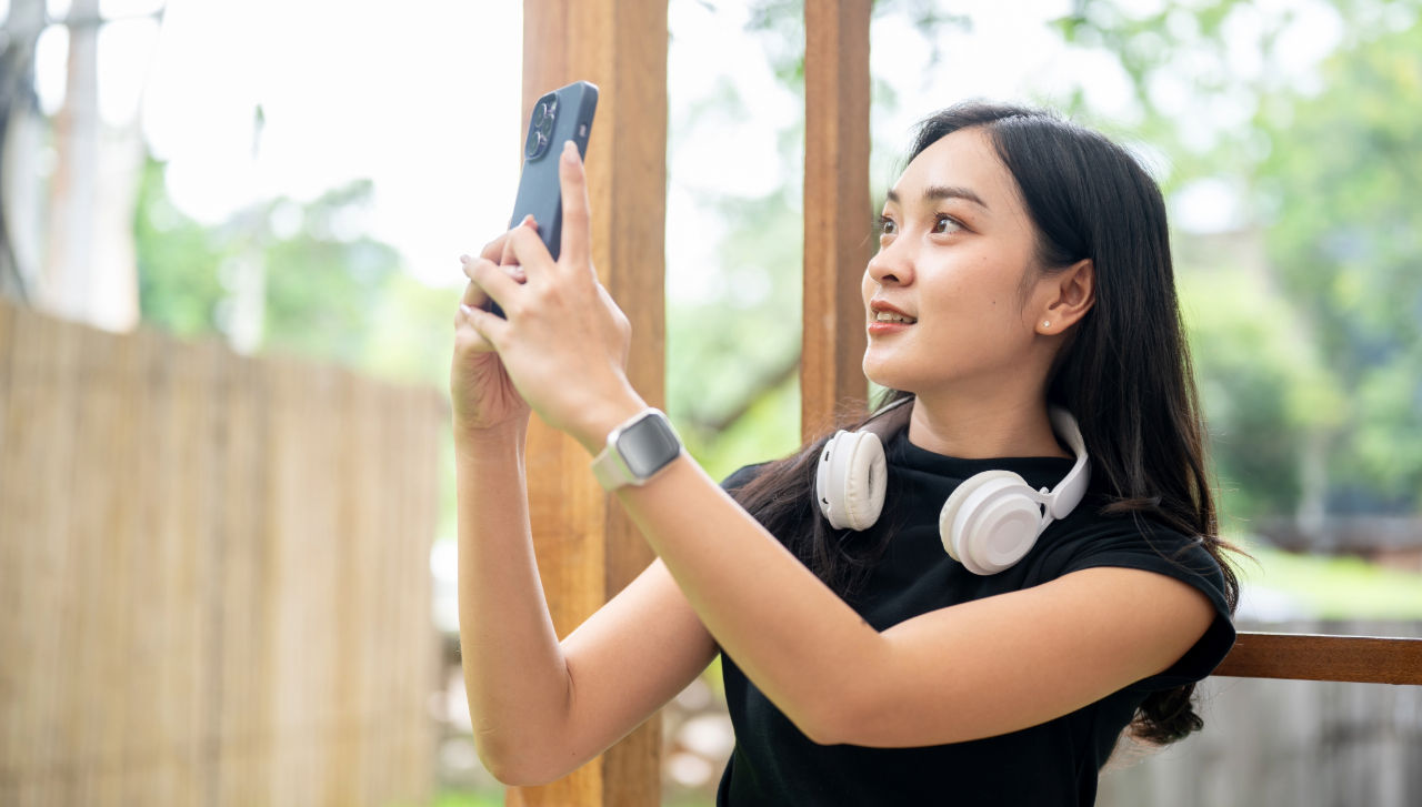 Isang babaeng may hawak na smartphone para kumuha ng litrato ng mga posibleng isyu sa kaligtasan na hindi pang-emergency, habang nakatayo sa labas sa isang natural na kapaligiran. Nakasuot siya ng smartwatch at may headphone na nakasabit sa leeg niya. Tampok sa background ang mga istrukturang gawa sa kahoy at mga halaman.